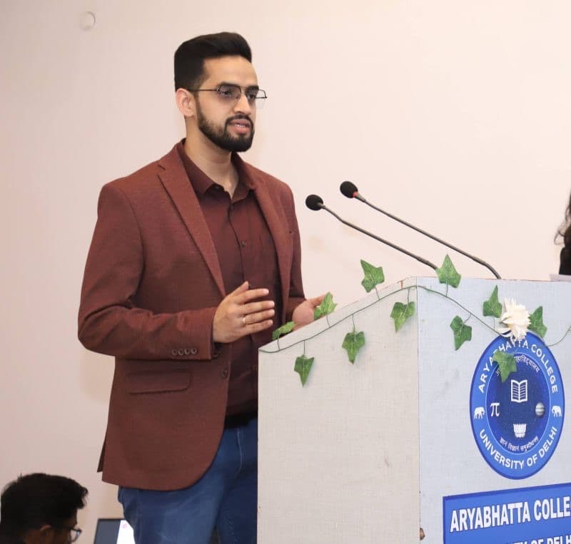 Speaking at a lectern marked Aryabhatta College, University of Delhi, microphones angled in.