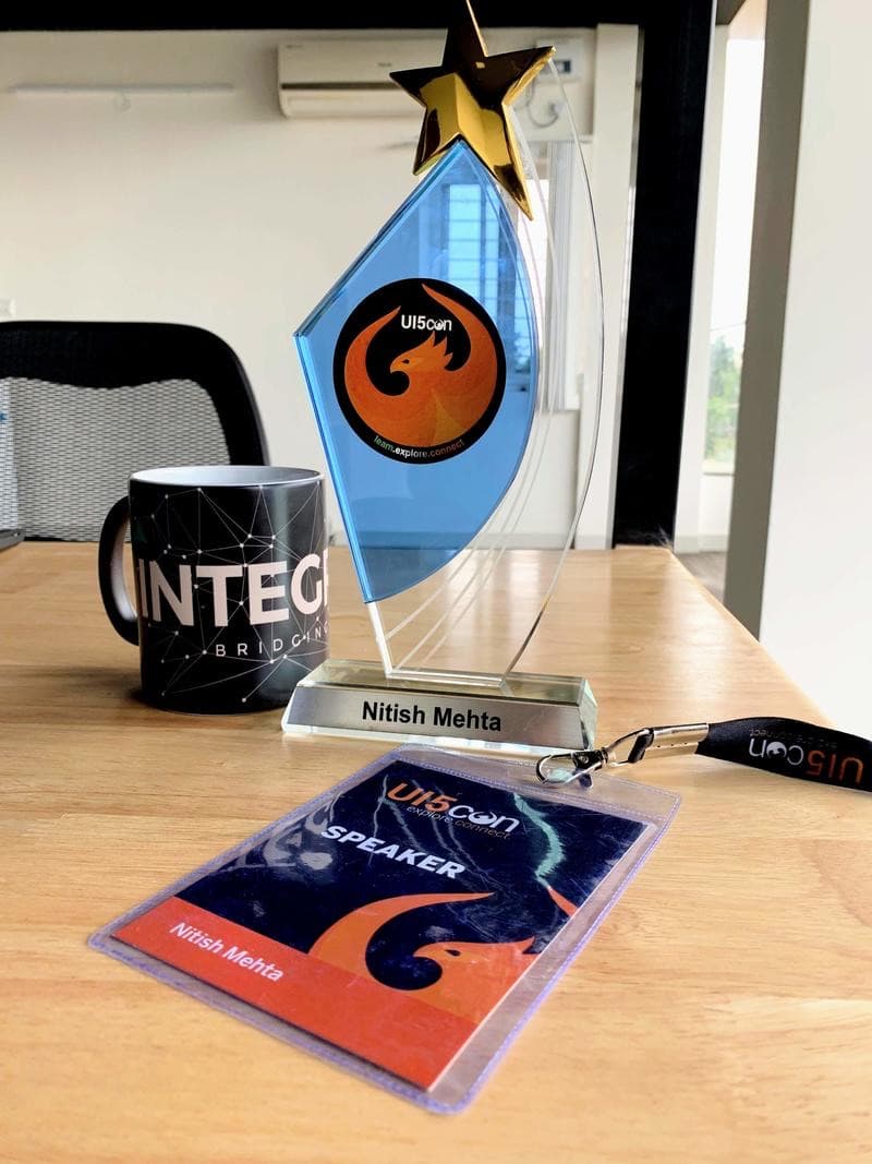 A UI5con trophy engraved Nitish Mehta on a desk, next to a speaker badge on a lanyard and an INTEGRTR mug.