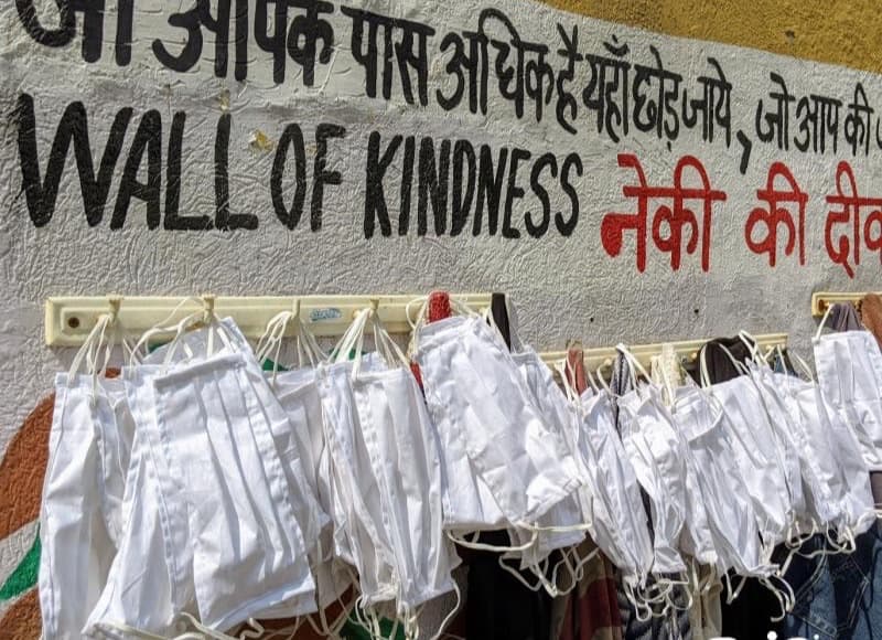 Rows of white cloth masks hanging from a rail under a street wall painted Wall of Kindness in English and Hindi.